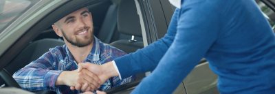 Menager leaning on front door and shaking hands with client after purchase. Young man in blue sweater helping his friend choosing vehicle. Brunette driver sitting in grey car and looking at brother.