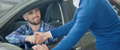 Menager leaning on front door and shaking hands with client after purchase. Young man in blue sweater helping his friend choosing vehicle. Brunette driver sitting in grey car and looking at brother.