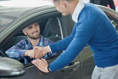 Menager leaning on front door and shaking hands with client after purchase. Young man in blue sweater helping his friend choosing vehicle. Brunette driver sitting in grey car and looking at brother.