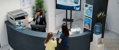 Receptionist working in hospital waiting area and offering support to patients at checkup visit, mother with child having medical appointment with physician. Reception desk at healthcare clinic.